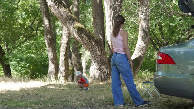 A peaceful mother and her curious child relax near their car in a sunny forest clearing. They feel happy and content spending quiet family time together.