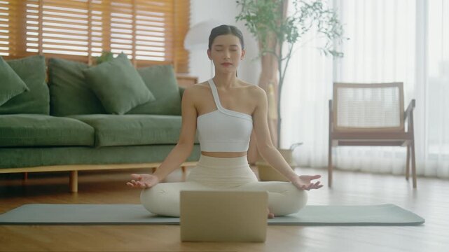 Young Asian woman meditating in lotus position at home, sitting on the floor in fitness clothing. Breathing workout to achieve relaxation, peace and mindfulness