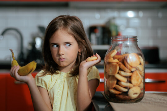 Little Girl Comparing Cookie And Banana Thinking About Healthy Snack Options. Thoughtful young girl holds cookie and banana deciding which healthy snack to choose