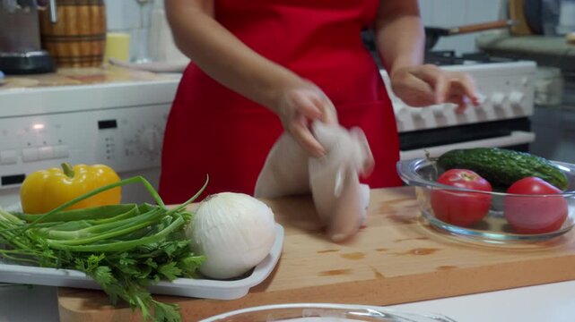 person in red apron meticulously wipes chicken on a wooden cutting board in a well-lit kitchen, surrounded by fresh vegetables and herbs, creating a clean and neutral atmosphere for food.