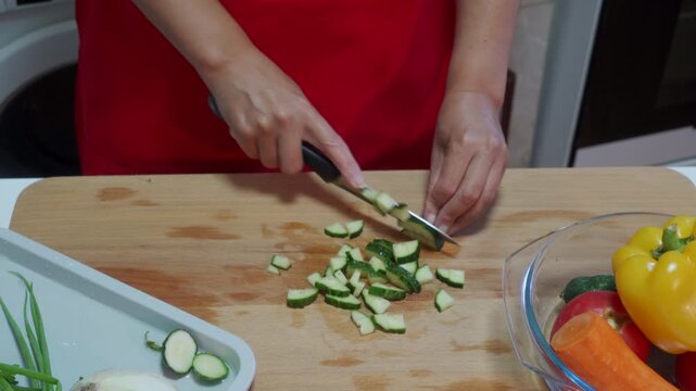 Woman in a red shirt is carefully chopping zucchini on a wooden cutting board in an overhead kitchen setting, emphasizing a focused and healthy eating tutorial for cooking enthusiasts.
