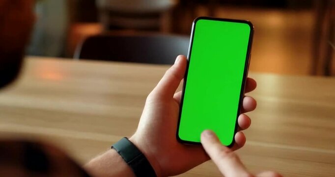 Person using smartphone with green screen mockup while sitting at wooden table indoors.