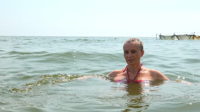 Mature woman stands chest deep on a shallow sandbar in green sea water, looking at the camera, with a fishing net on the hazy horizon