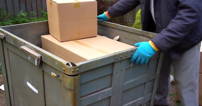 Person wearing blue gloves disposing of cardboard boxes into a large outdoor waste container.