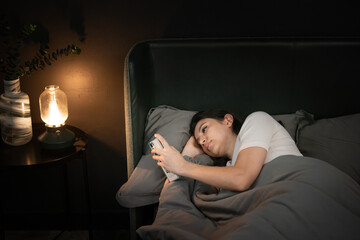 Fototapeta premium Lying in bed woman with insomnia scrolls her phone under warm lamp light creating quiet restless mood in dim bedroom scene