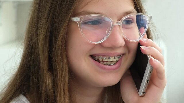 Close up teenage girl with dental braces and glasses smiling talking on mobile phone. Orthodontic treatment and vision care concept