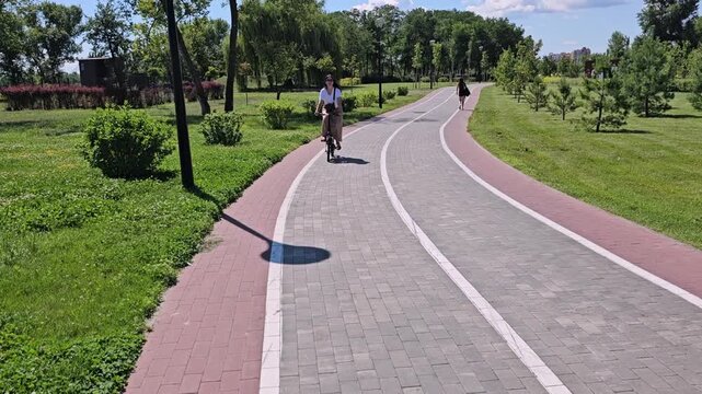 Wide shot of a woman in sunglasses and casual attire riding a small black folding electric bicycle along a paved bike path in a sunny green park.