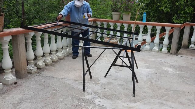 Young Man Applying Black Paint with Brush to Metal Bed Frame on Terrace