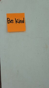 Hand Sticking Orange Sticky Note with Phrase Be Kind on Wall