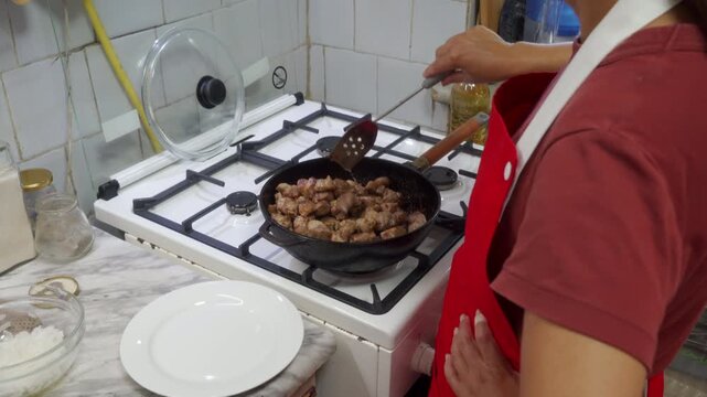 A person wearing a red apron is stirring food in a pan on a gas stove in a cozy kitchen with tiled walls and a glass bowl nearby, creating a homely and focused cooking scene with warm.