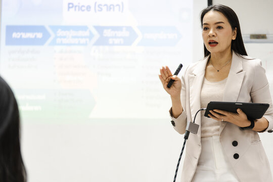 Woman presents confidently with tablet and marker. She gestures while speaking, engaged and professional. Presentation slide shows price details in Thai script. Her focused expression suggests clarity