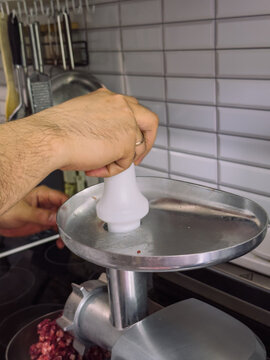 Hand using pusher to feed raw meat into electric meat grinder during homemade minced meat preparation in kitchen.