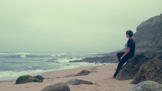 Young man sits on a  big stone on the empty seashore, cloudy rainy weather.  Sad young guy feels bad and melancholy, and looks into the ocean distance. Thoughtful human feel loneliness and depression