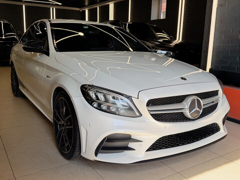 Front  view of a white Mercedes-AMG C 43 luxury sports sedan parked in a detailing studio. Modern premium German car with LED headlight, radiator grille with brand logo, black alloy wheel. 