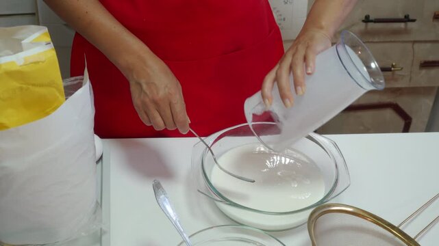 A focused baker in a red apron pours fresh milk into a glass bowl on a white table. They mix ingredients with joyful, calm care. The bright kitchen feels cozy and comforting.