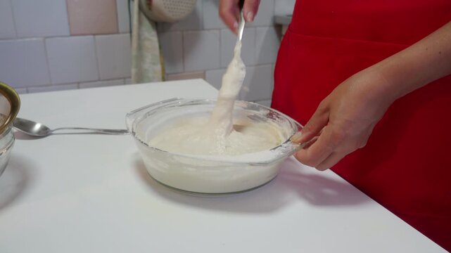 A focused baker in a red apron stirs smooth dough inside a glass bowl on a white table. They create the recipe with joyful, calm care. The bright kitchen feels warm and cozy.