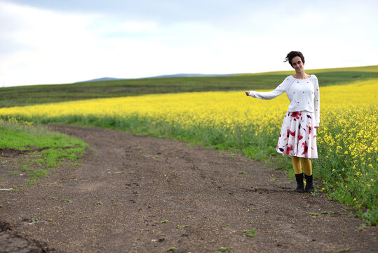 Woman hitchhiking on a dirt road next to a yellow canola flower field
