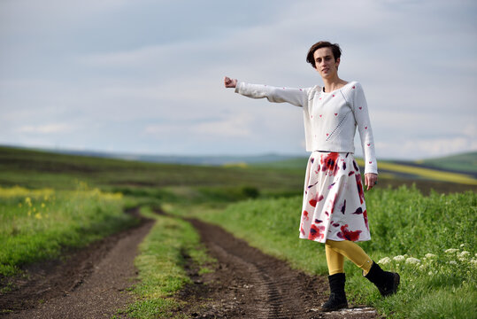 Woman hitchhiking on a dirt road next to a yellow canola flower field