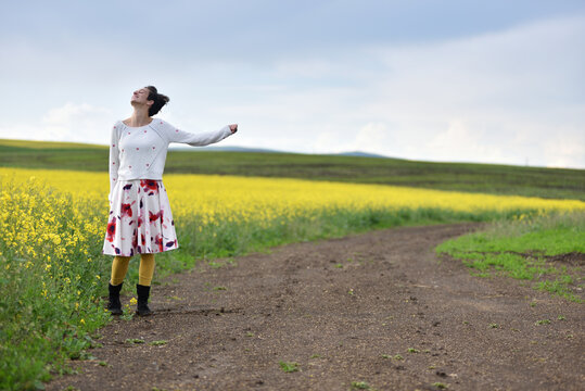 Woman hitchhiking on a dirt road next to a yellow canola flower field