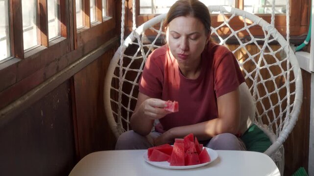 A content woman eats fresh watermelon slices while sitting in a hanging chair on a sunny balcony. She feels relaxed and refreshed enjoying a sweet, peaceful summer snack.
