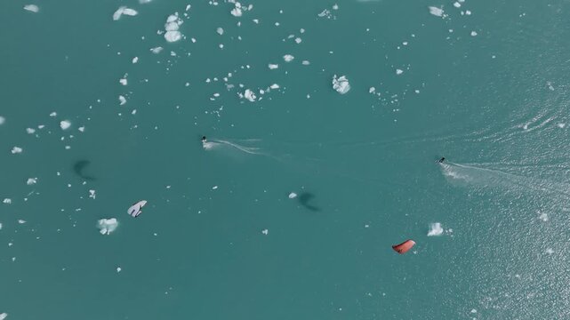 Top-down drone tracks kitesurfers weaving through floating ice on Lago Argentino near El Calafate, Santa Cruz, Argentina, beneath turquoise glacial waters, drone top-down tracking shot.
