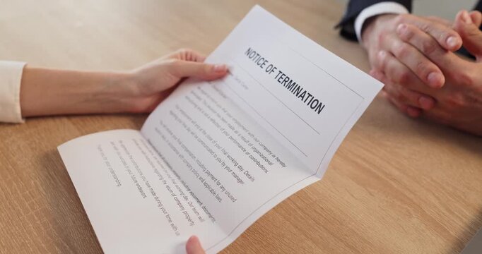Employee reads termination notice during manager meeting. Close view shows a dismissal document held across a desk in a modern office. Concept shows professional job loss and uncertainty.