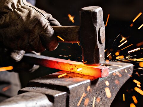 A close-up shot of a blacksmith striking a glowing hot metal bar on an anvil with a heavy hammer.