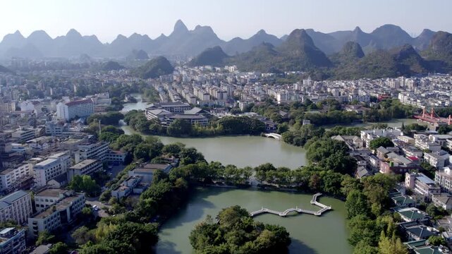 A panoramic aerial view of a city with interconnected lakes, surrounded by distinctive karst mountains under clear sky.