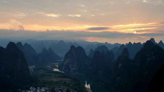 A serene landscape of steep limestone karst peaks surrounding a winding river, captured at sunrise with soft warm light in the sky.