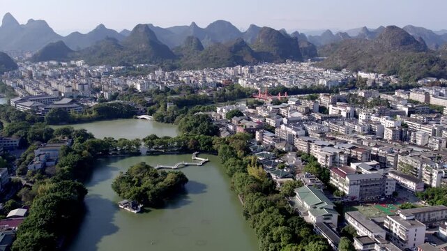 A panoramic aerial view of a city surrounded by distinctive karst mountains, with lakes and urban buildings interspersed with greenery.
