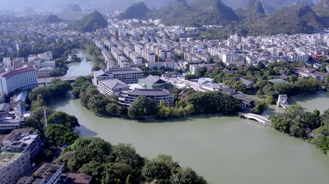 An aerial shot shows an urban area with a winding river, surrounded by karst mountains and lush vegetation under clear skies.