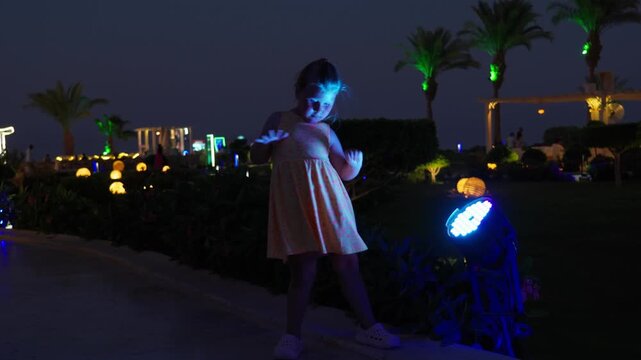 A little girl emotionally dances in front of a bright street lamp at night. Happy energetic kid on summer vacation at a resort.