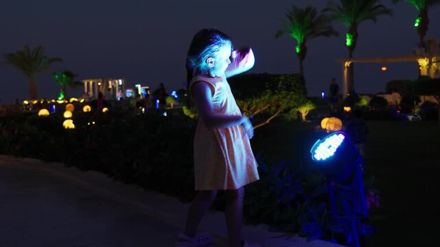 A little girl emotionally dances in front of a bright street lamp at night. Happy energetic kid on summer vacation at a resort.