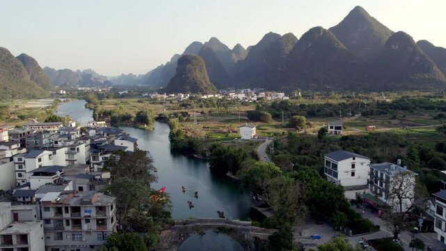 A scenic rural landscape featuring an old stone arch bridge spanning a river, flanked by riverside buildings and backed by karst mountains.