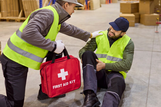 Warehouse workers handle injury accident emergency. Colleague kneels with first aid kit evaluates leg pain and follows safety protocol in a busy warehouse. Concept safety response in workplace.