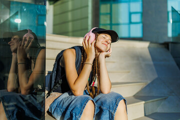 Fototapeta premium Zoomers, Relaxed teenager listening to music sitting near glass building.