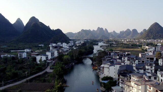 A serene rural landscape featuring a river winding through a village, flanked by karst mountains under a pale sky.