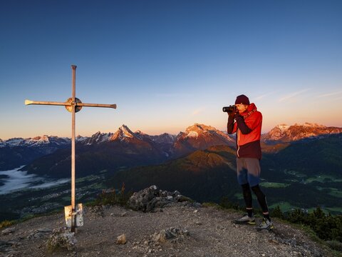 Photographer at Rauher Kopf summit cross at sunrise, in the background the Berchtesgaden Alps, Steinernes Meer, Sch&ouml;nfeldspitze, Watzmann, Hochkalter and horse-rider Alpe, Berchtesgaden National Park, Bischofswiesen, Berchtesgadener Land, Upper Bavaria, Bavaria, Germany