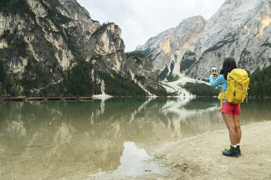 Woman hiker taking smartphone photo of Lago di Braies alpine lake turquoise water mountain reflections Dolomites Italy