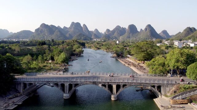 A wide river flows under a multi-arch bridge, backed by iconic karst mountains with lush green vegetation along the banks.