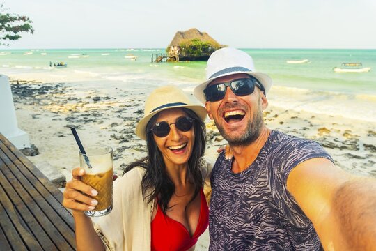 Happy couple smiles at camera takes playful selfie on a tropical beach with famous attraction rock restaurant behind. Authentic joy, laughter, cheers holiday maker and summer vacation vibes in paradise