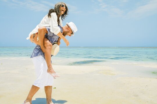 Multi-ethnic happy couple having fun piggyback riding on white sand beach. Caucasian man and middle aged woman in playful moment on the shore at summer time - Concept of happiness and travel
