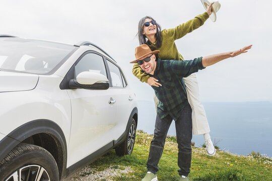 Close up happy couple enjoys holiday time together piggyback ride by their white SUV during Mediterranean road trip. Lifestyle shot capturing love, freedom adventure on scenic coastal cliff side