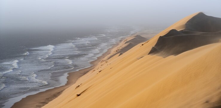 Sand dunes with wave patterns and sea in evening light, wind blowing sand across the dunes, Atlantic coast and sandy desert with sea of dunes, impressive desert landscape, Sandwich Harbour, Namib Naukluft Park, Namib Desert, Namibia