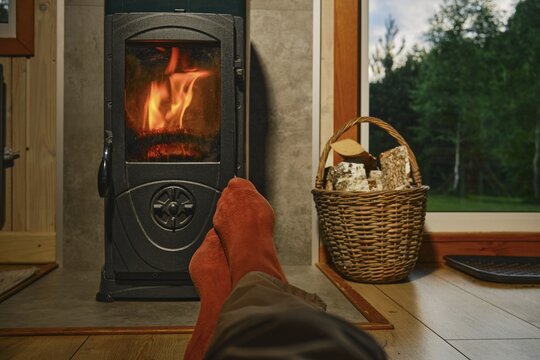Feet in red socks are resting in front of wood stove in wooden cabin. Flames are visible, creating warmth. A basket with firewood sits nearby. Cabin interior is simple with wooden floor and large window