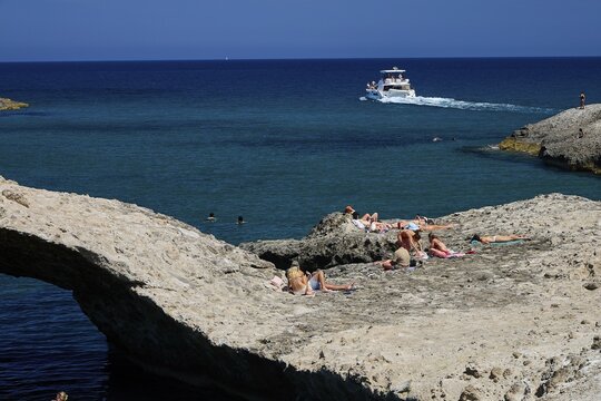 People sunbathe on rocky water's edge while a boat passes by, Kapras, Kapros, Beach, Kapros Beach, Caves, Paralia Kapros, Milos, Cyclades, Greek Islands, Greece