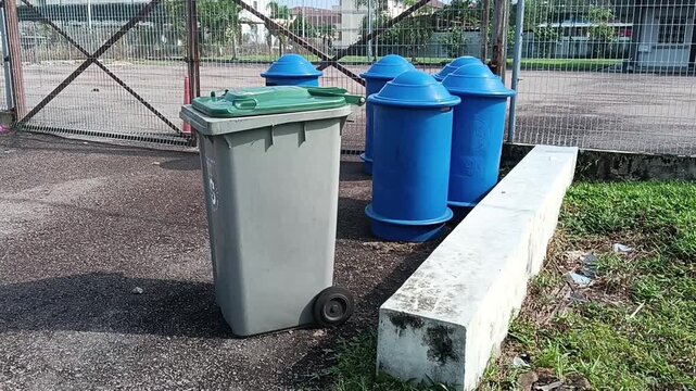 A gray wheeled garbage bin with a green lid stands on a paved area beside several blue outdoor waste bins near a fenced compound. The image captures a clean waste disposal area in an urban setting wit