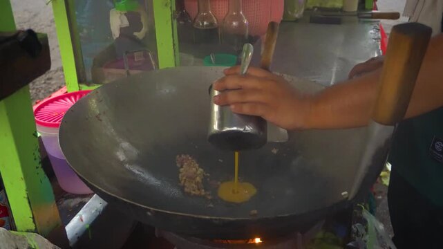 A local street food vendor pouring egg into a wok with sauteed shallots and garlic at a traditional stall, preparing a delicious fried rice dish.