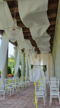 Elegant wedding ceremony decor with white drapes, crystal chandeliers, and chiavari chairs swaying in the wind in a stone colonnade with a floral arch