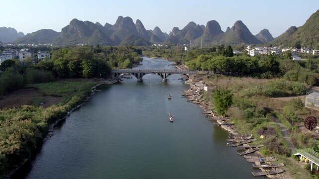 A river with a stone bridge, flanked by villages and trees, backed by distinctive karst mountain peaks.